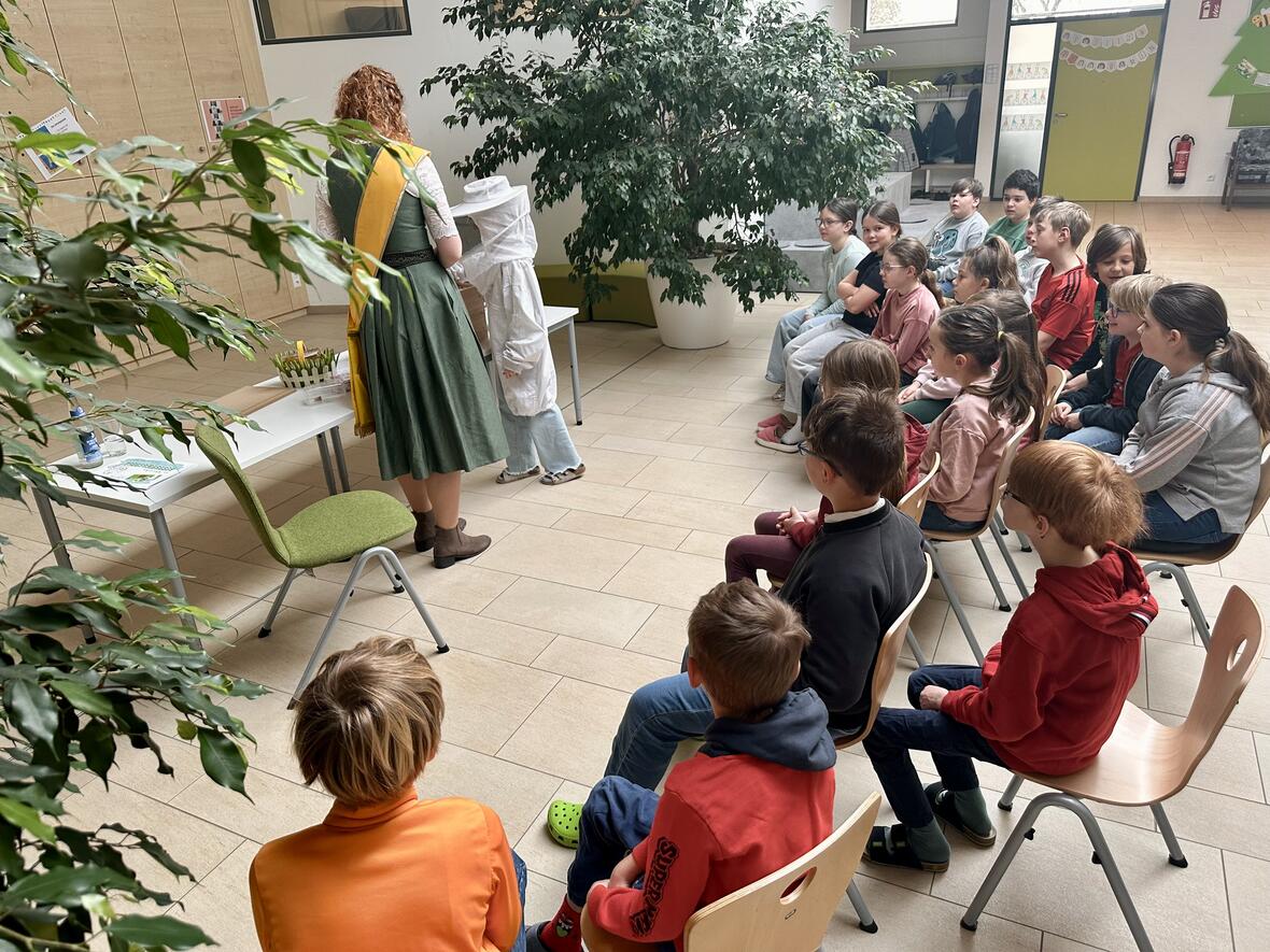 Kinder sitzen in einem Vorführraum, während eine Frau mit Bienenschutzkleidung erklärt.