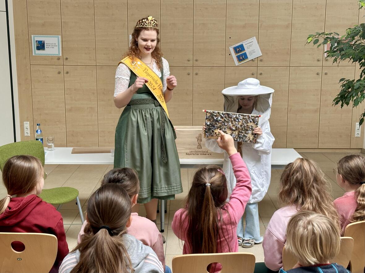 Bayerische Königin erklärt Kindergruppe das Thema Bienenzucht, während eine Schülerin ein Bienenvölker-Bild zeigt.