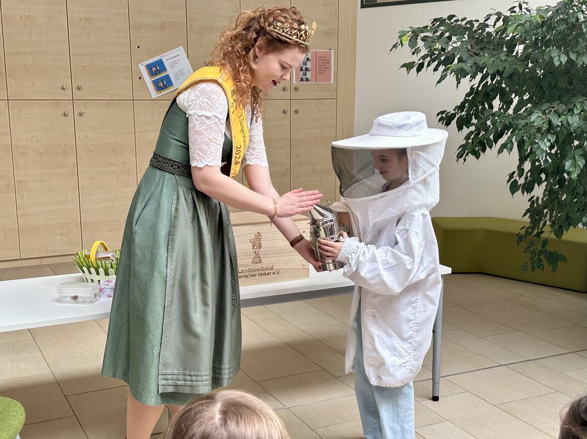 Eine Bienenkönigin überreicht einem Kind im Schutzanzug einen Smoker. Hintergrund mit Pflanzen und Tischen.