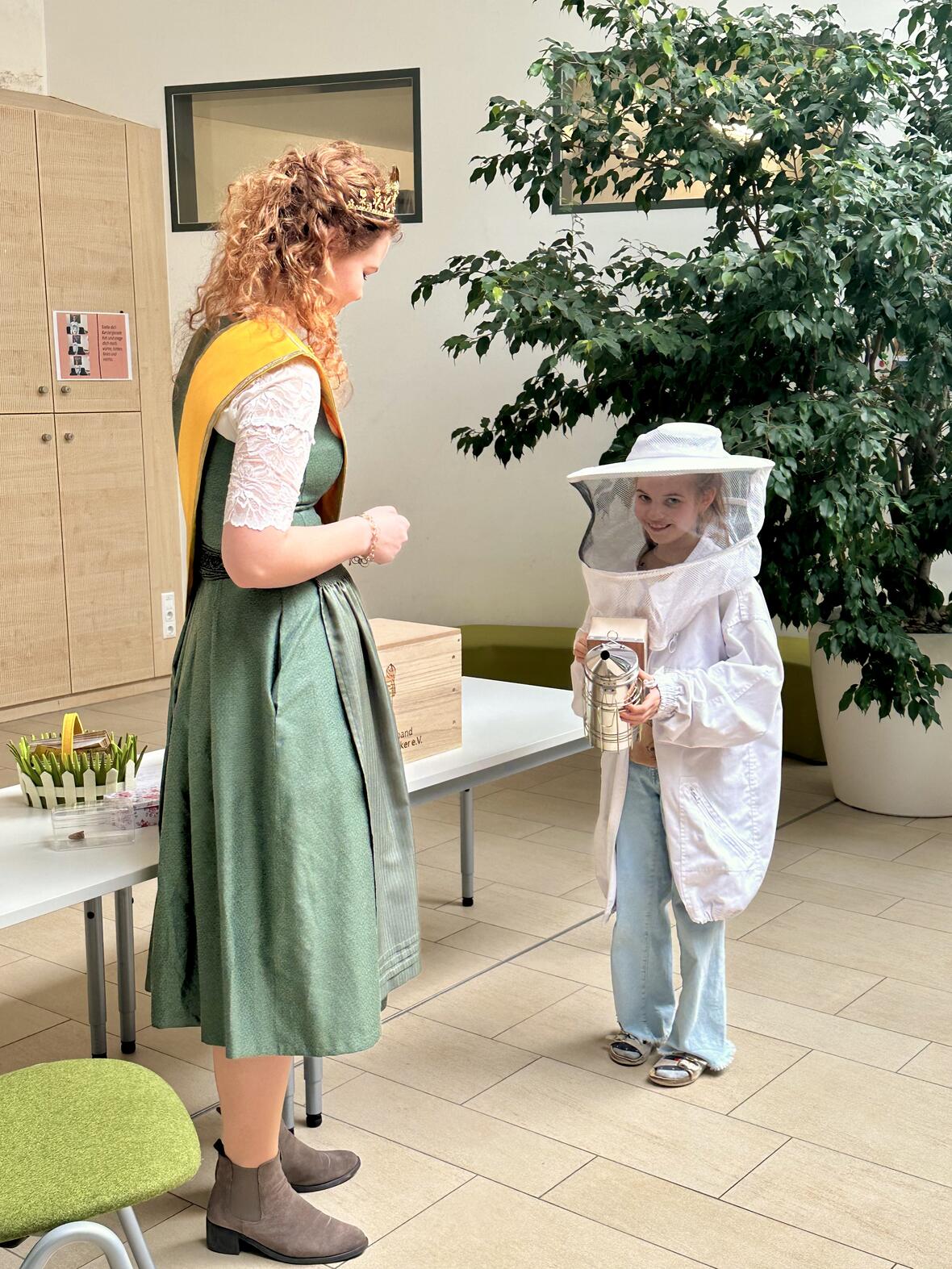 Kind im Bienenanzug lächelt eine Dame mit Krone und Tracht an, im Hintergrund ein Tisch mit Bienenutensilien.