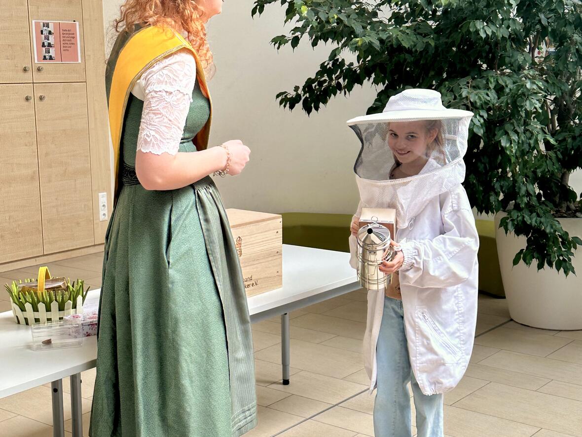 Kind im Bienenanzug lächelt eine Dame mit Krone und Tracht an, im Hintergrund ein Tisch mit Bienenutensilien.