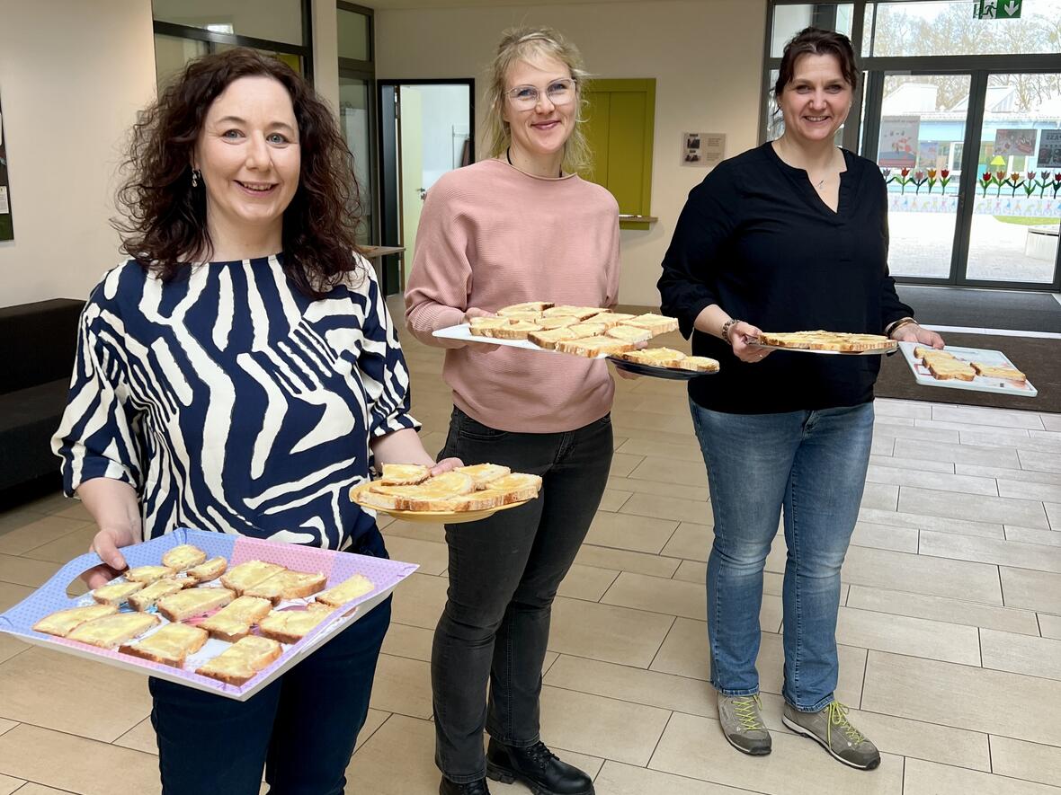 Drei Frauen in einem hellen Raum halten Tabletts mit belegten Broten.