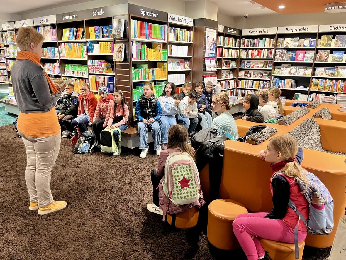 Eine Person erklärt etwas vor einer Gruppe von Kindern in einer Bibliothek. Bücherregale im Hintergrund.