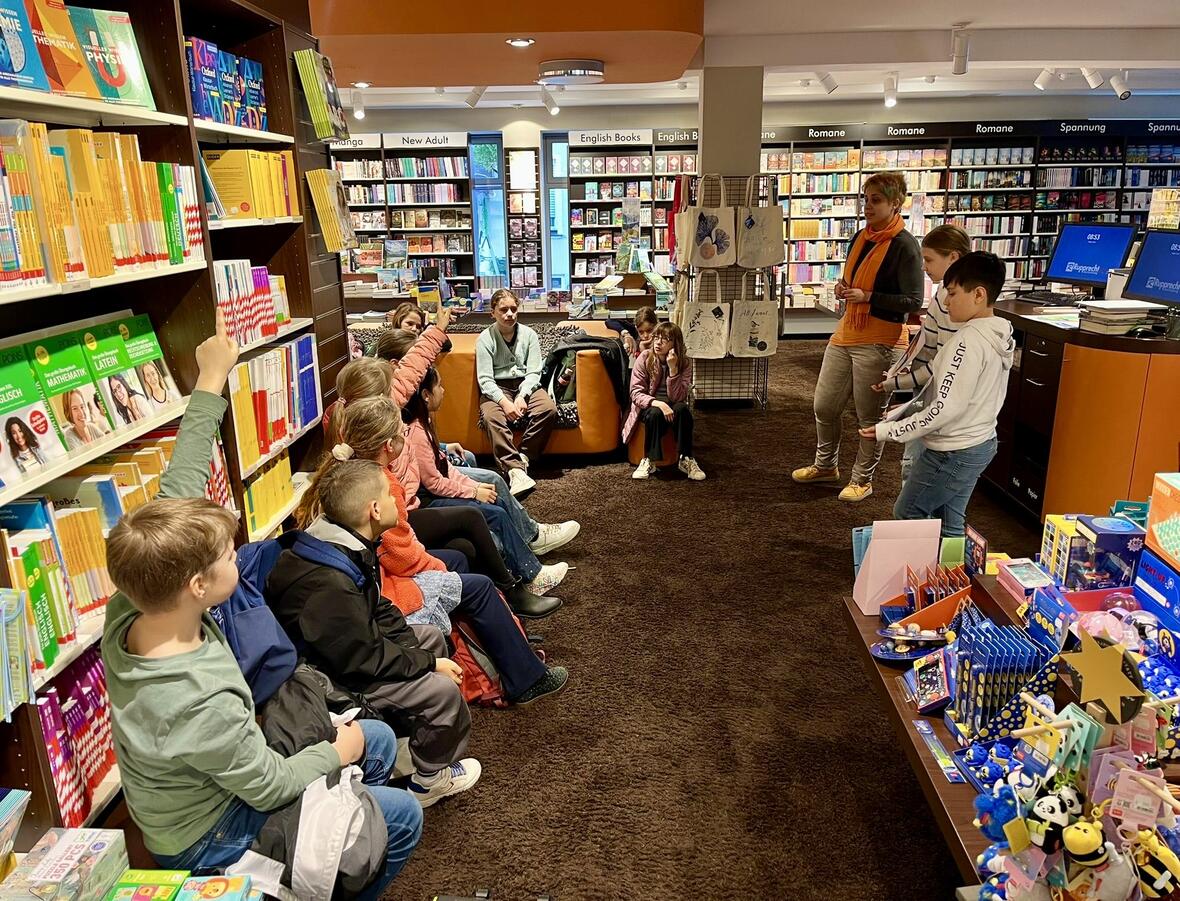Kinder sitzen in einer Buchhandlung und hören der Referentin mit orangefarbener Schal zu. 