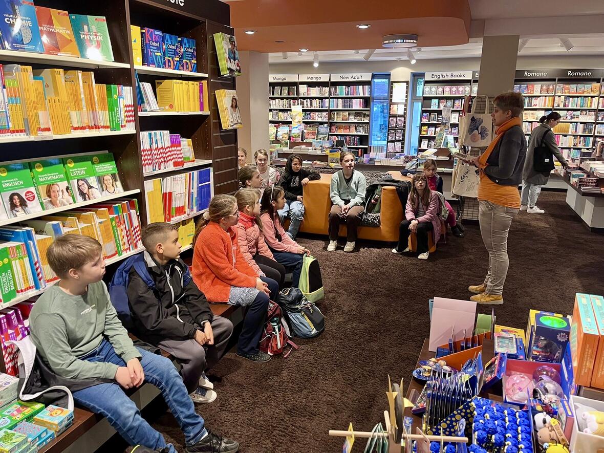 Gruppe von Kindern hört einer Vortragenden in einer Buchhandlung zu, umgeben von Regalen mit Schulbüchern.