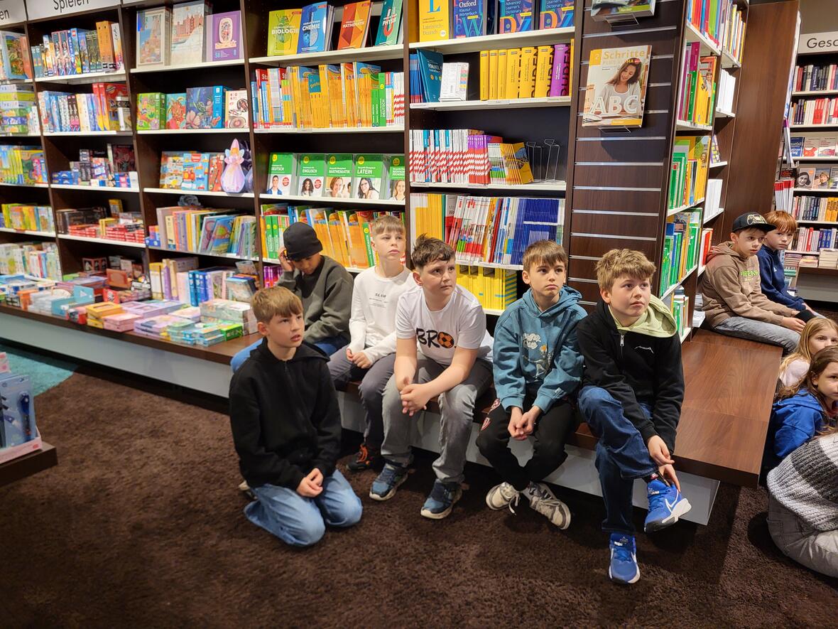 Sechs Kinder sitzen im Buchladen auf dem Teppich und hören gebannt zu. Bücherregale im Hintergrund.