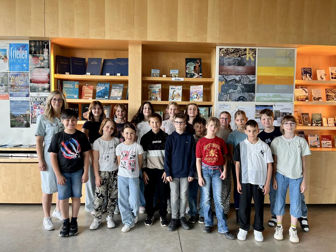 Gruppe von Schülern und einer Lehrerin in einer Bibliothek, umgeben von Büchern und Informationsmaterialien.