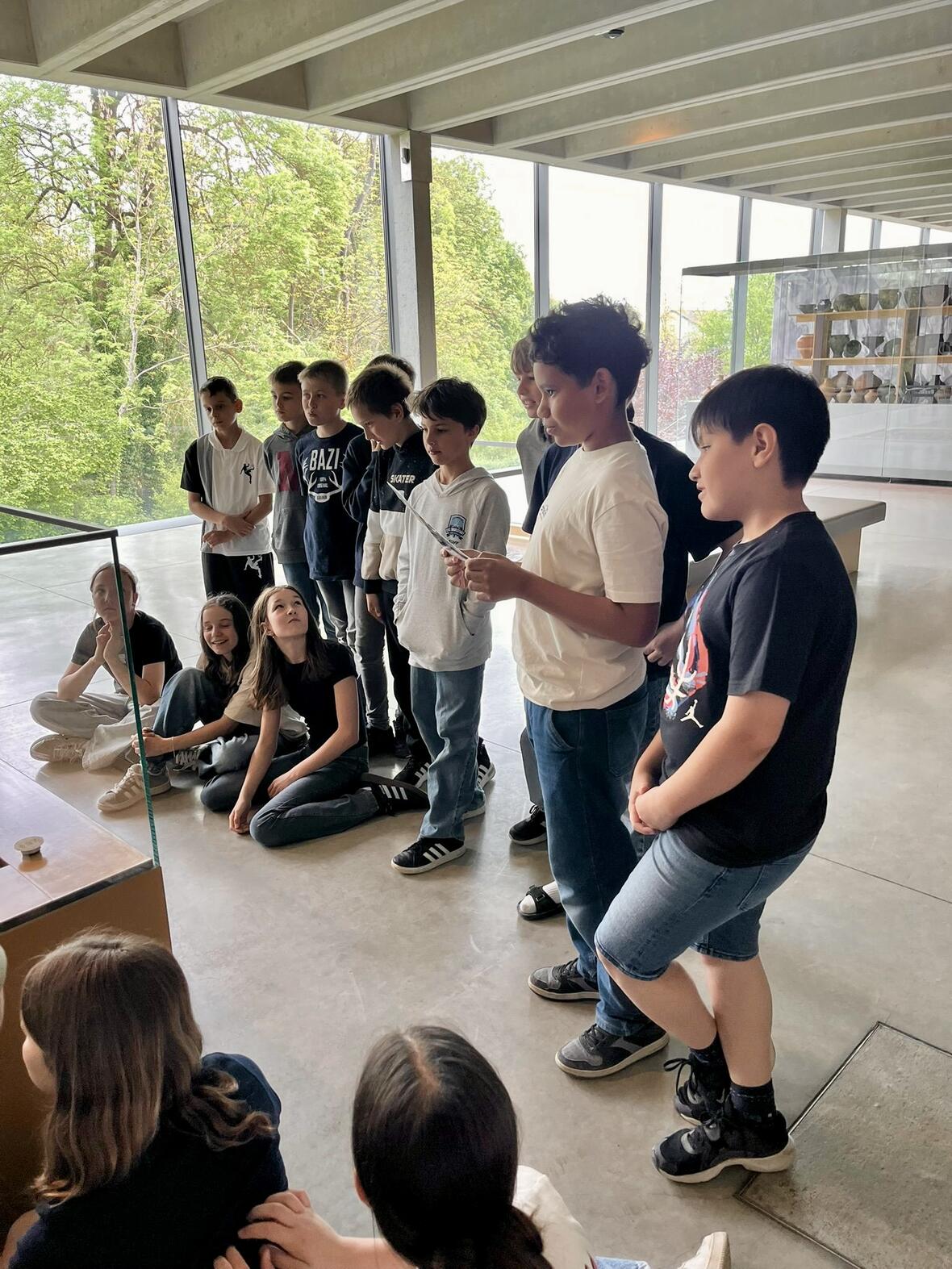 Schülergruppe hört einem Mitschüler zu, während sie im Ausstellungsraum vor großen Fenstern stehen.
