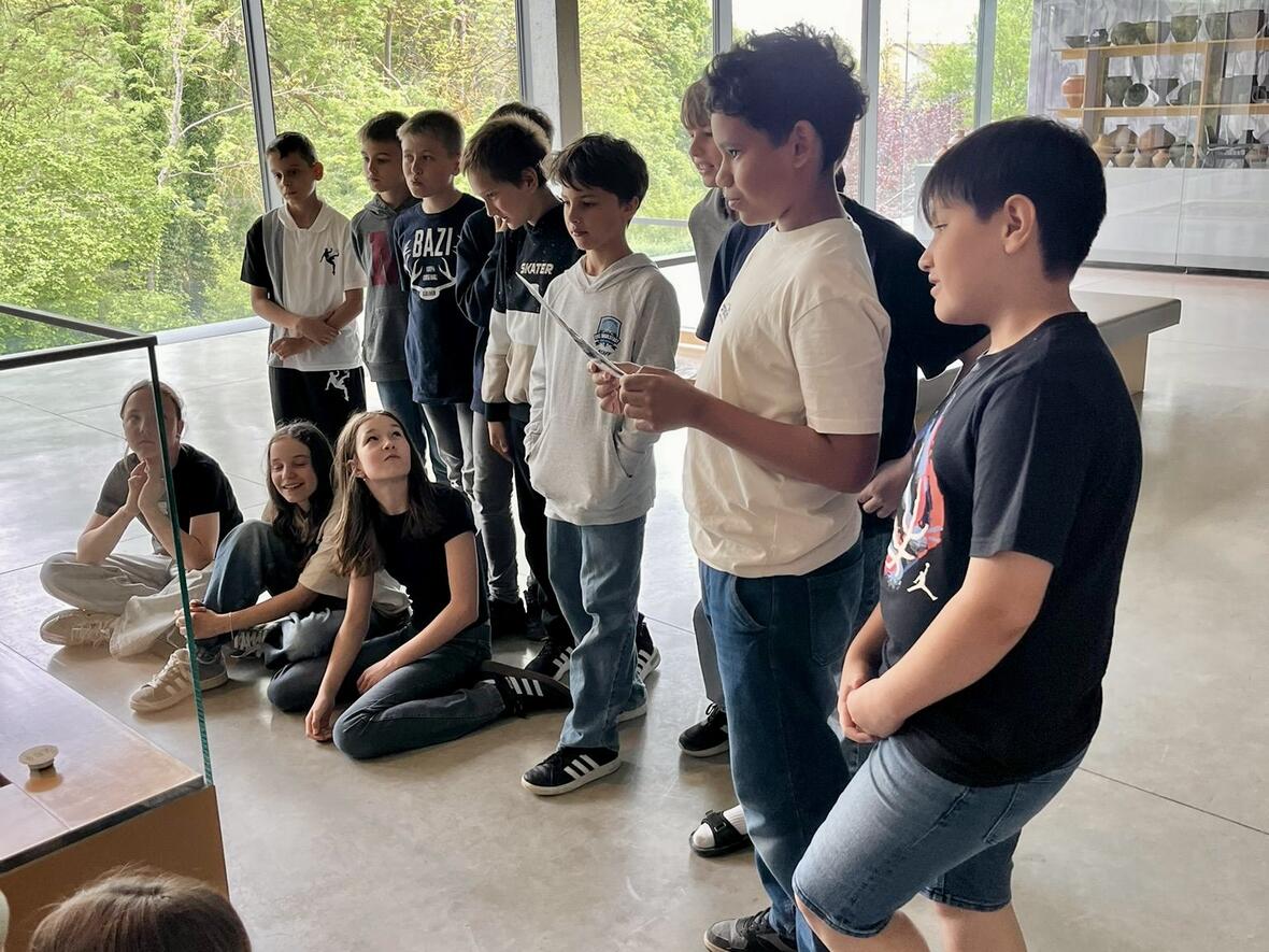 Schülergruppe hört einem Mitschüler zu, während sie im Ausstellungsraum vor großen Fenstern stehen.