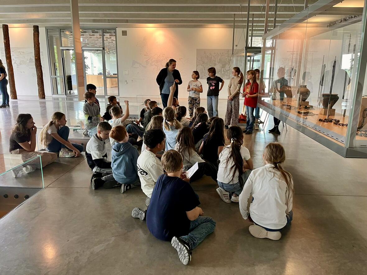Schulgruppe sitzt auf dem Boden in einem Museum, während eine Lehrerin ein Exponat präsentiert.