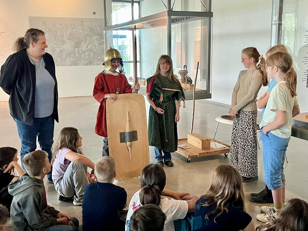 Kinderexperimente im Museum: Ein Junge im römischen Kostüm erklärt mit Schild, während die Gruppe zuhört.