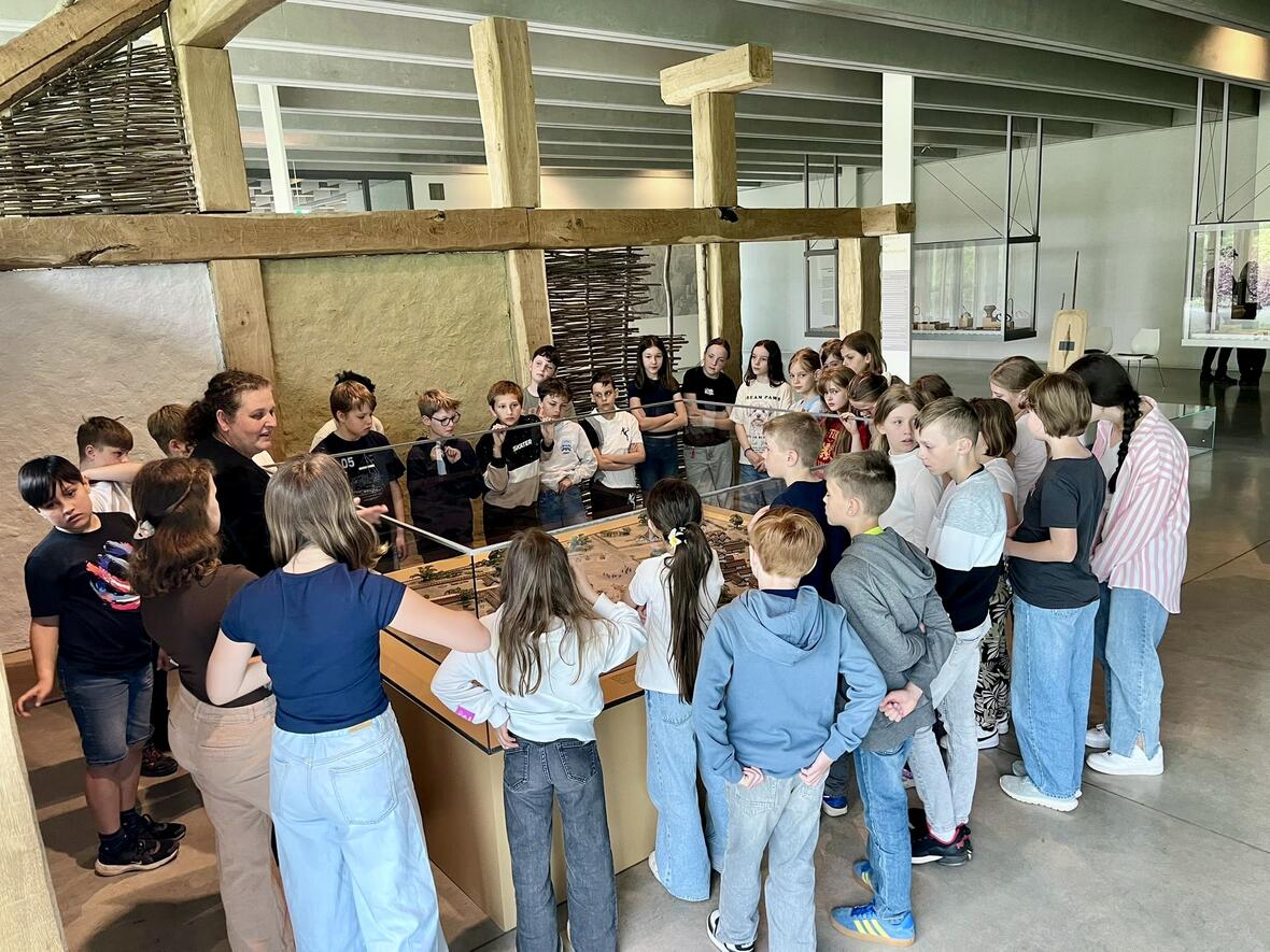 Schülergruppe hört einer Lehrerin im Museum aufmerksam zu und betrachtet ein Diorama.