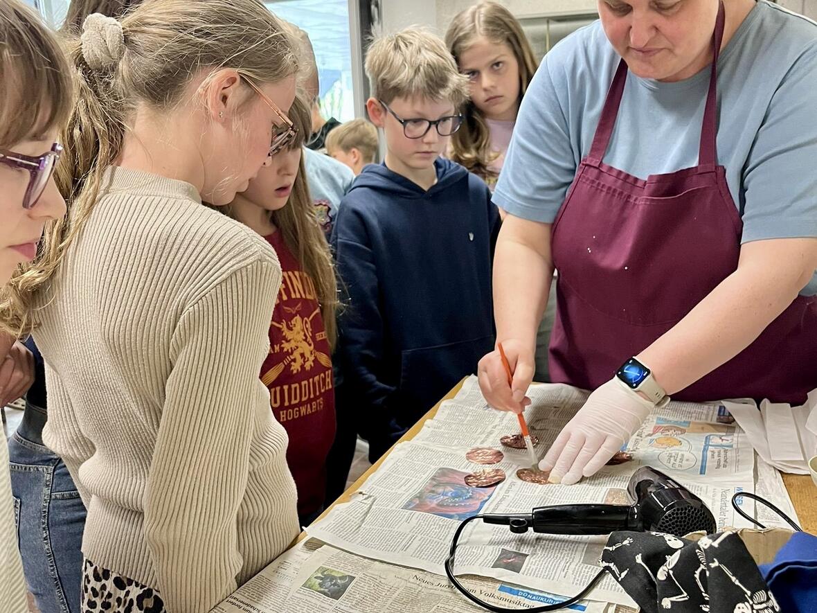 Schüler*innen beobachten eine Lehrkraft beim Arbeiten mit Materialien auf Zeitungsblättern.