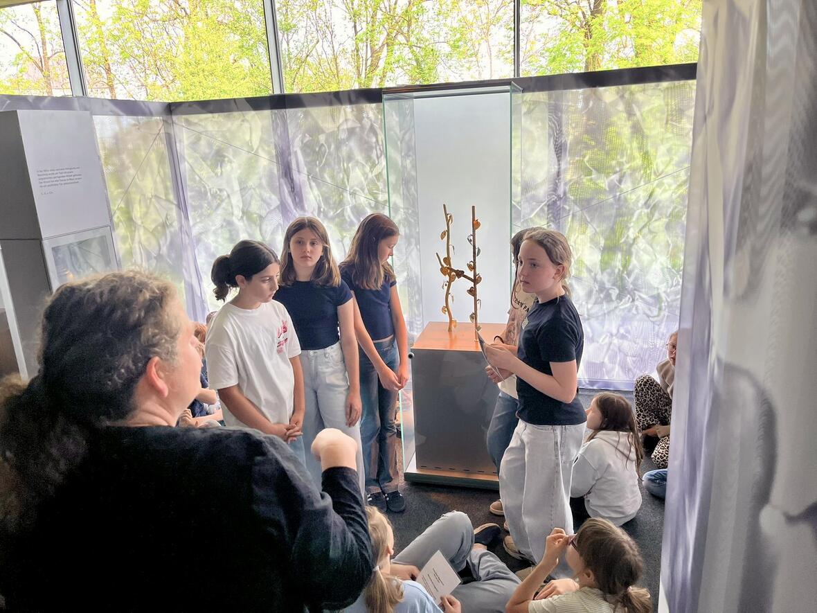 Gruppe von Kindern hört einer Erwachsenen bei einer Museumsführung aufmerksam zu.