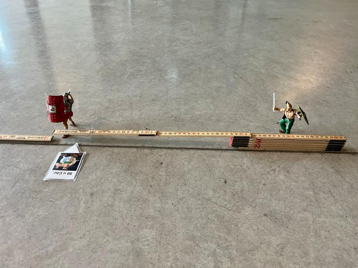 Zwei Figuren stehen auf einem Maßstab: ein Krieger mit Schild und ein anderer mit Schwert und Schild.