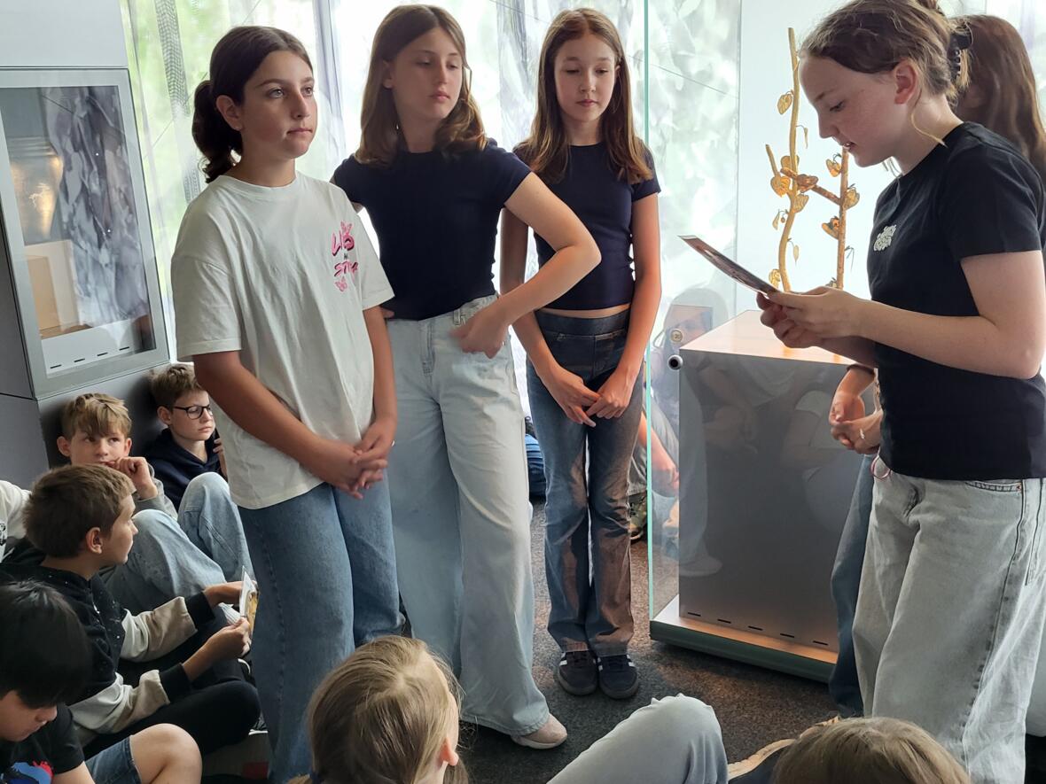 Mädchen diskutieren in einer Ausstellung, während andere Kinder auf dem Boden sitzen und zuhören.