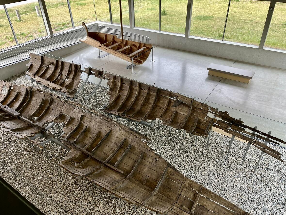 Ausgestellte Holzboot-Reste in einem modernen Ausstellungsraum, umgeben von Kiesboden.