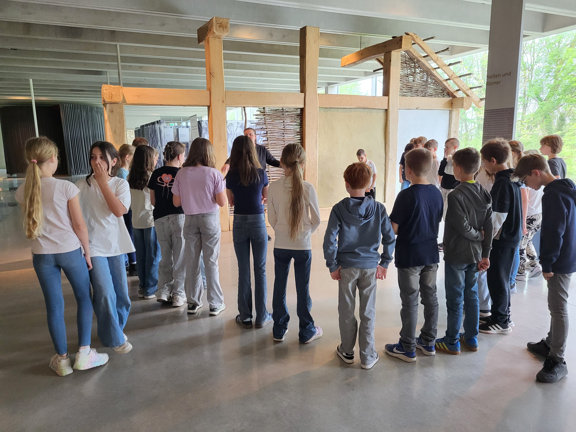 Schülergruppe im Museum hört einer Präsentation neben einem Nachbau eines historischen Gebäudes zu.