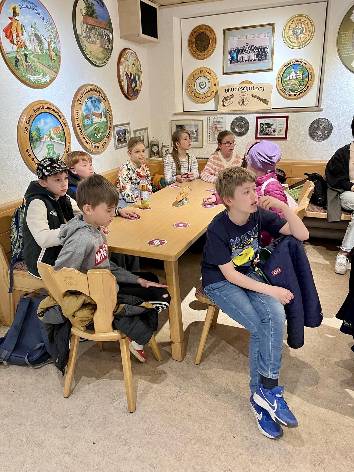 Gruppe von Kindern sitzt an einem Tisch in einem Raum mit Wanddekorationen und bespricht etwas.