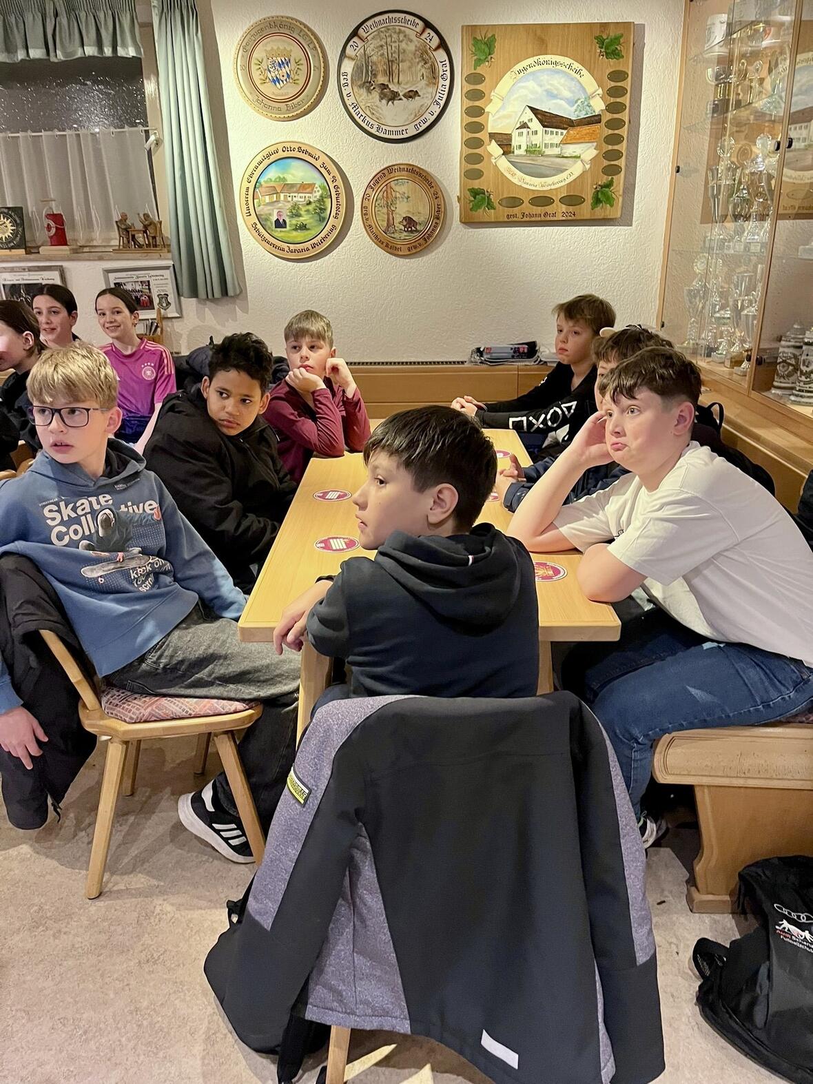  Gruppe von Kindern und Jugendlichen sitzt an einem Tisch in einem Vereinsraum, aufmerksam lauschend.