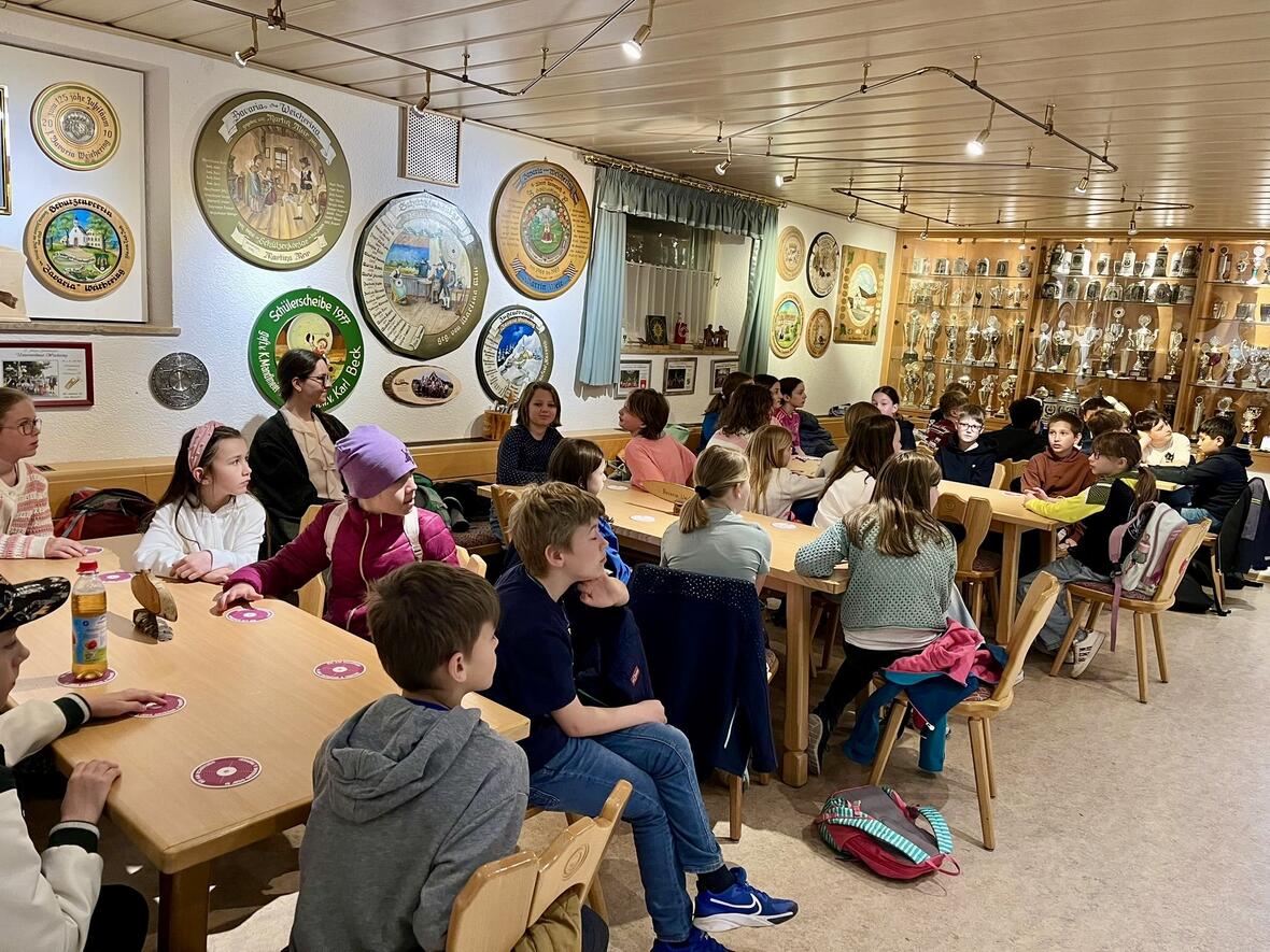 Gruppen von Kindern und Jugendlichen sitzen an Tischen in einem Vereinsheim, umgeben von Wappen und Pokalen.