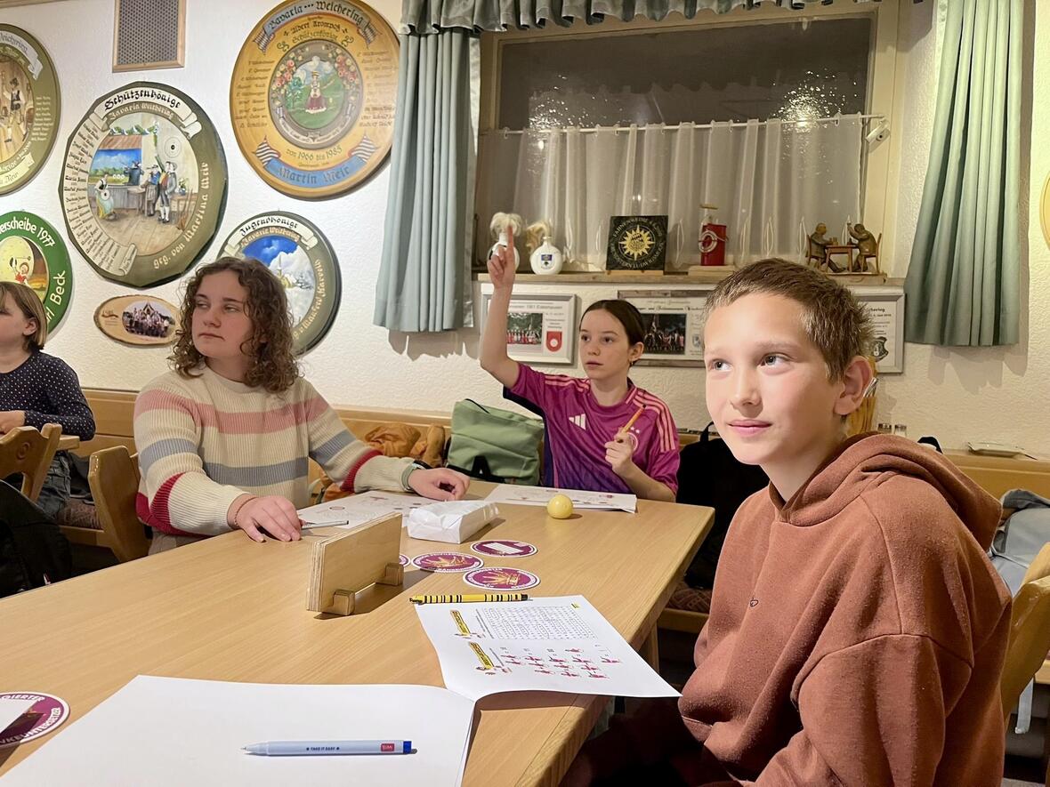 Drei Kinder in einem gemütlichen Raum mit Wänden voller Schilder, bei einer Spielfortsetzung am Tisch.