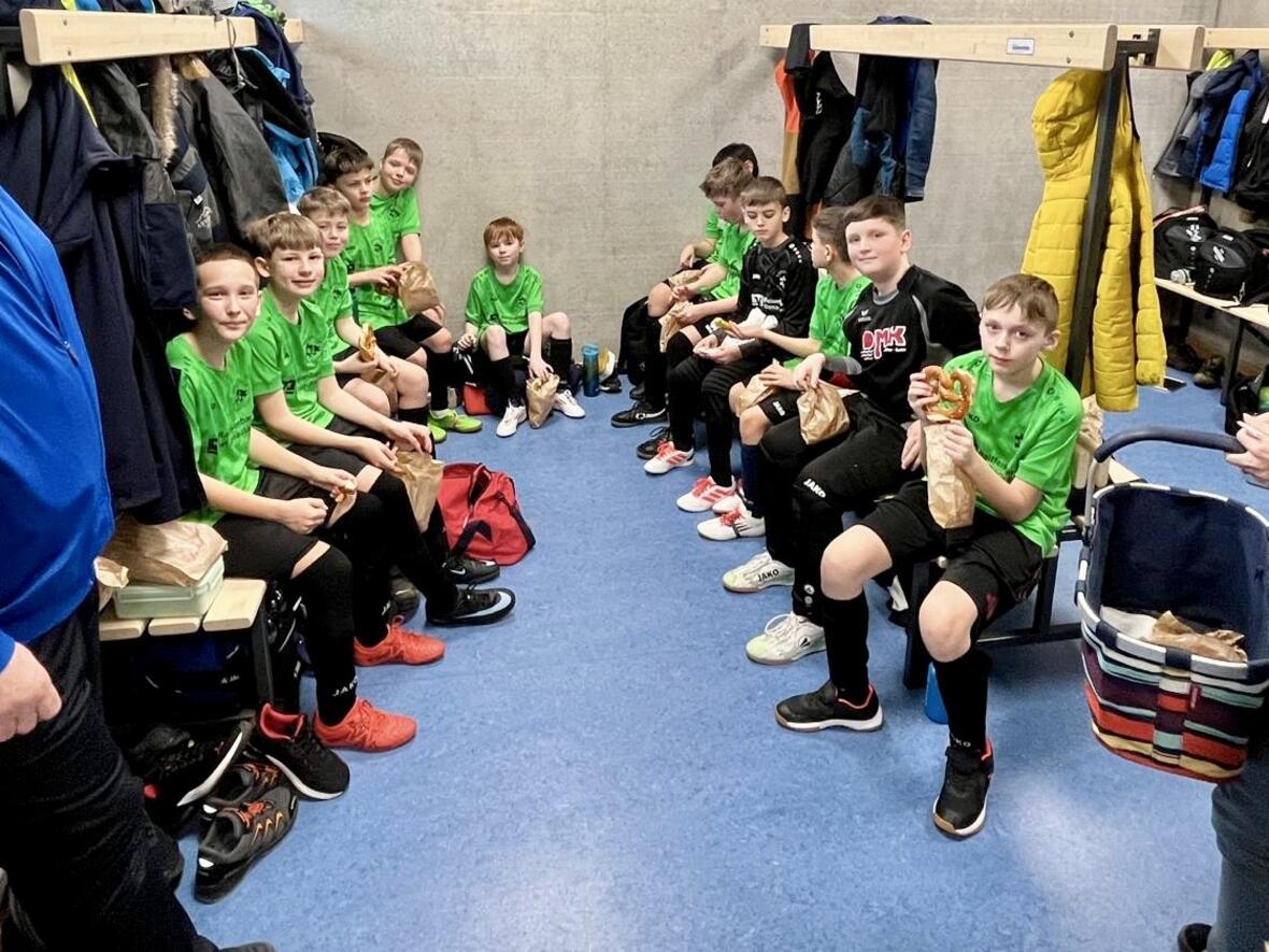 Gruppe von Fußballspielern in grünem Trikot sitzt in der Umkleide und isst Snacks.