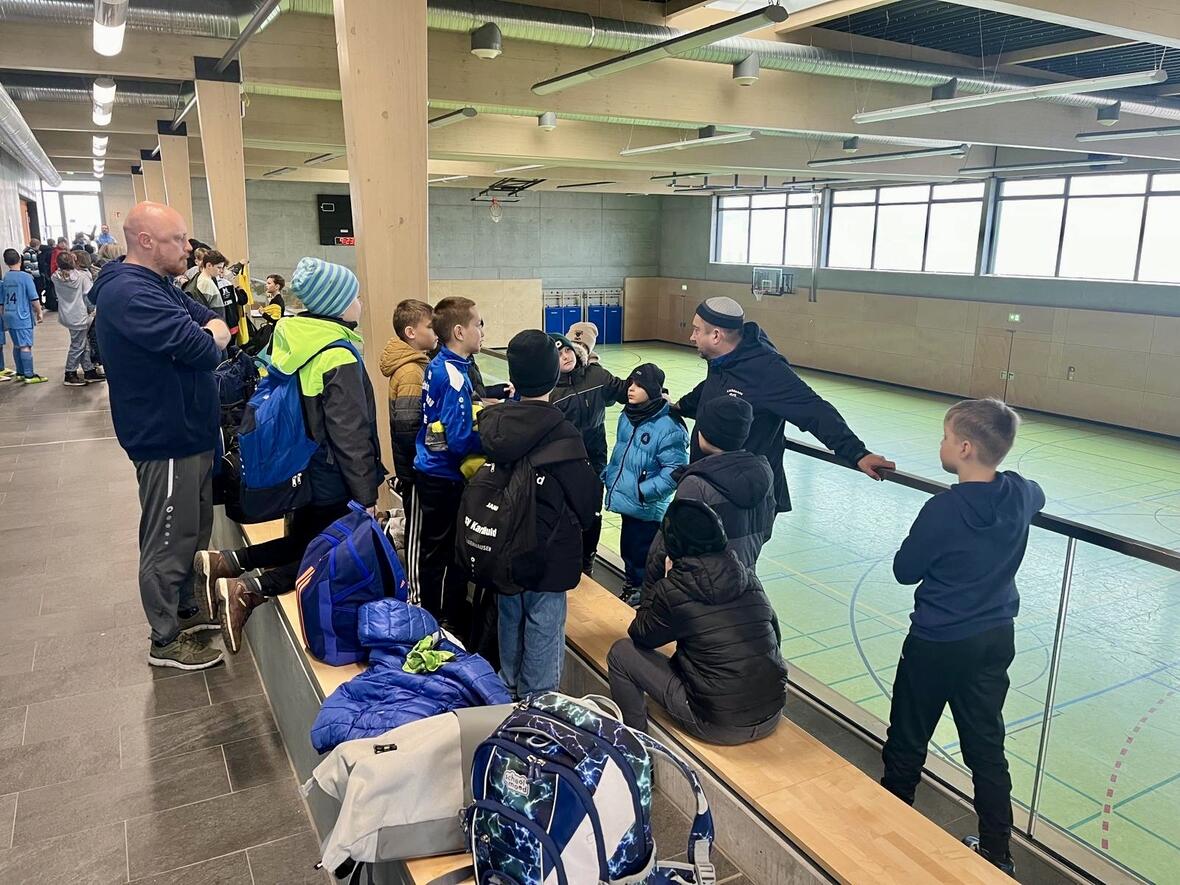 Gruppe von Kindern in Sportkleidung hört einem Trainer in einer Turnhalle zu. Taschen liegen auf der Bank.
