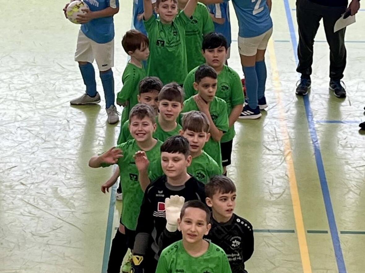 Kinderfußballmannschaft in grünen Trikots steht in der Halle, Trainer beobachtet sie.