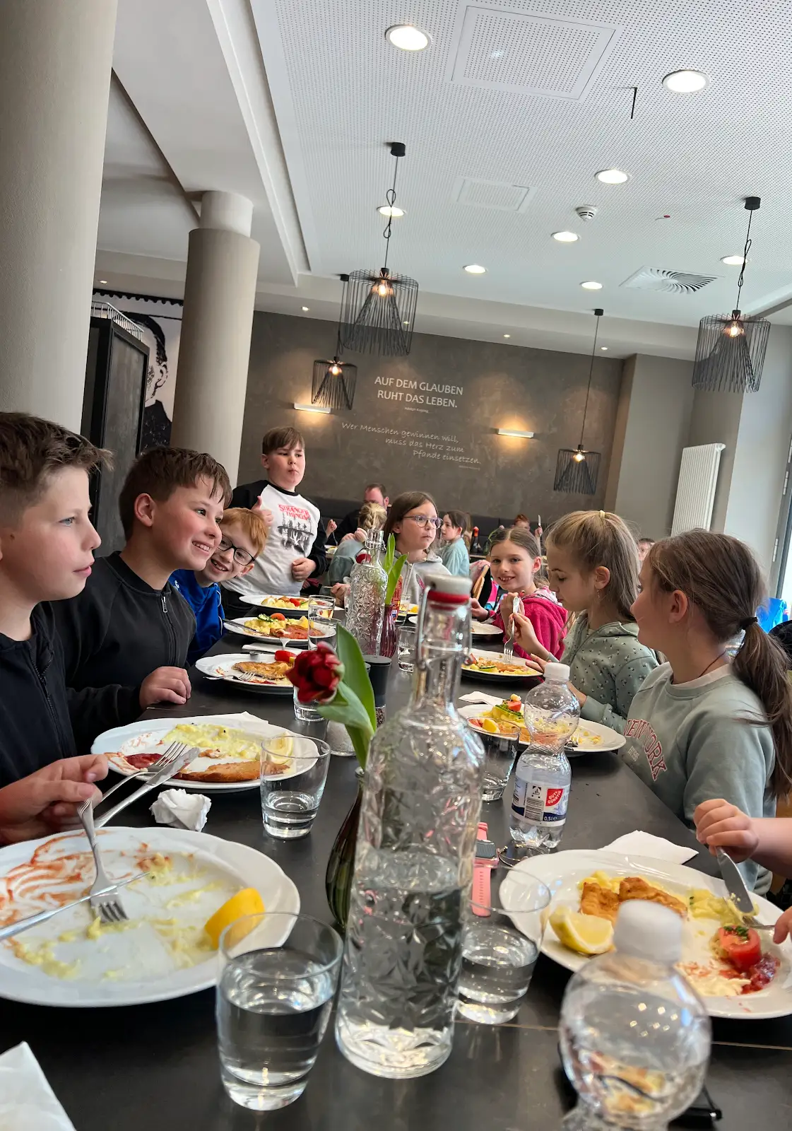 Kinder und Jugendliche essen gemeinsam in einem modernen Restaurant, geschmückt mit Blumen und Gläsern.