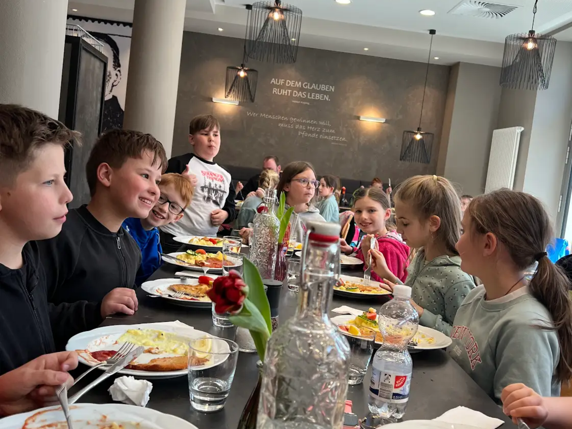 Kinder und Jugendliche essen gemeinsam in einem modernen Restaurant, geschmückt mit Blumen und Gläsern.