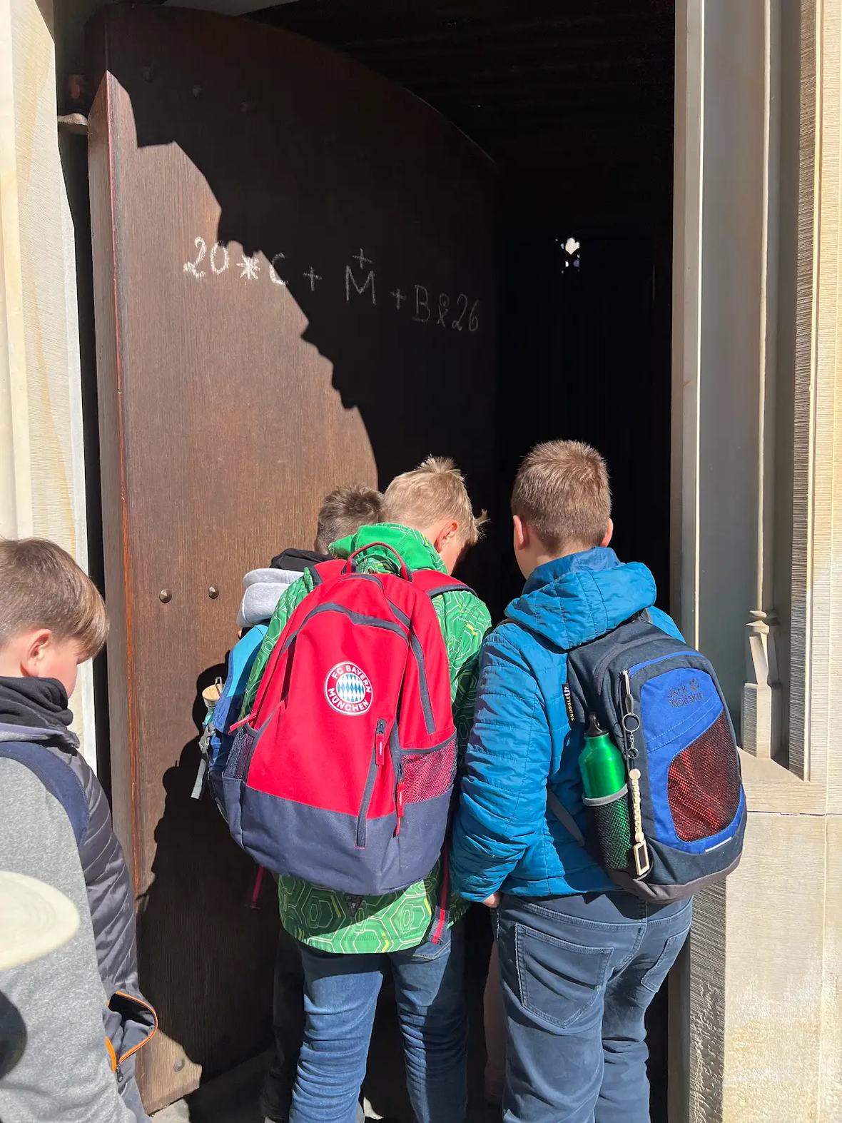 Gruppe von Kindern mit Rucksäcken, die ein großes, halboffenes Tor betrachten.