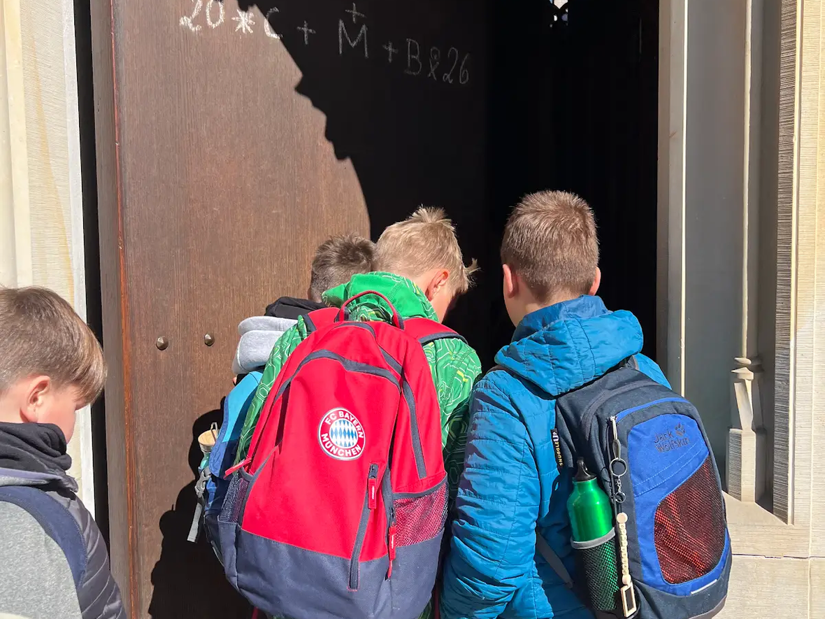 Gruppe von Kindern mit Rucksäcken, die ein großes, halboffenes Tor betrachten.