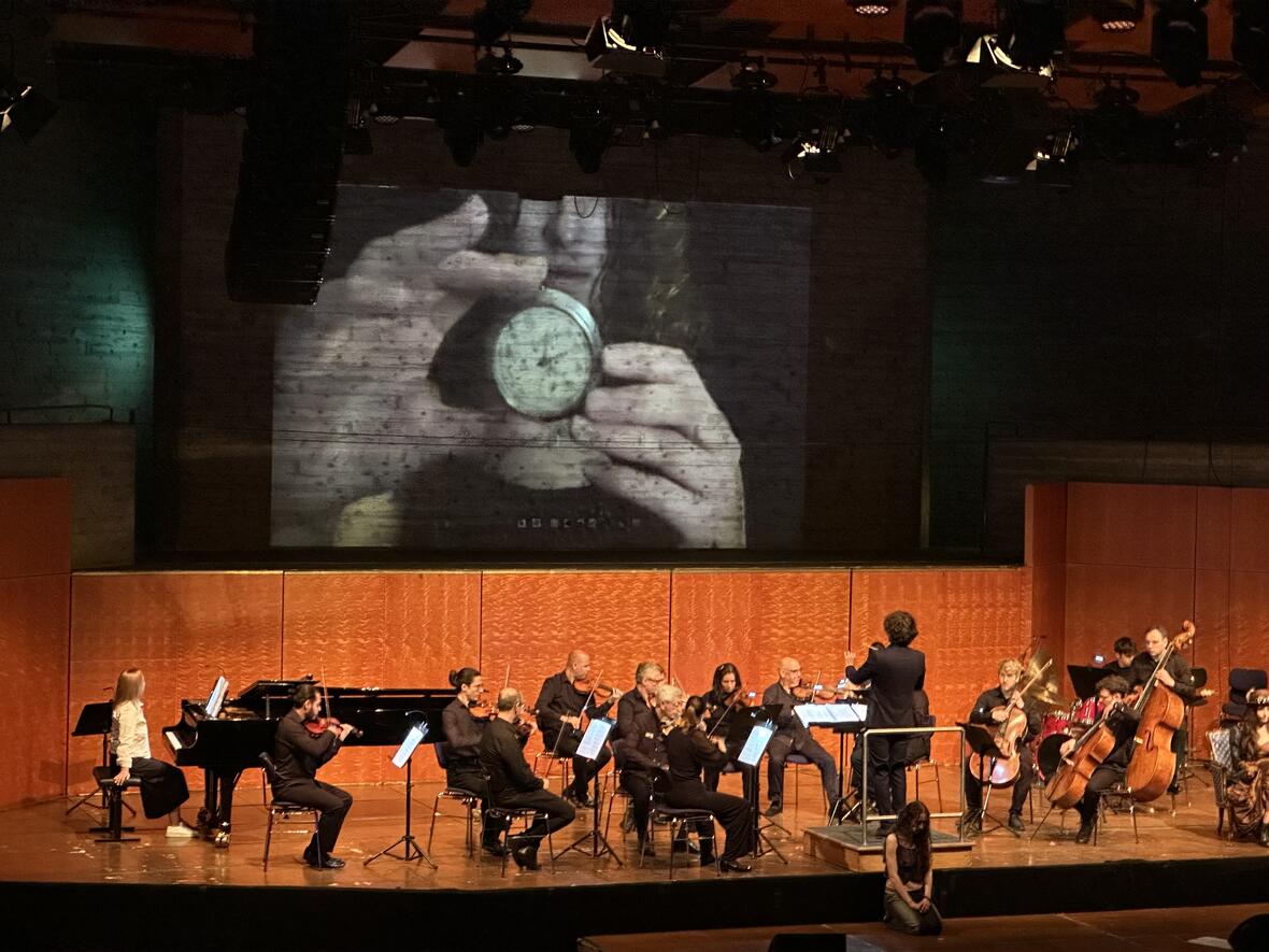 Konzert mit Orchester auf der Bühne, projizierte Nahaufnahme von Händen, die einen kleinen Gegenstand halten.