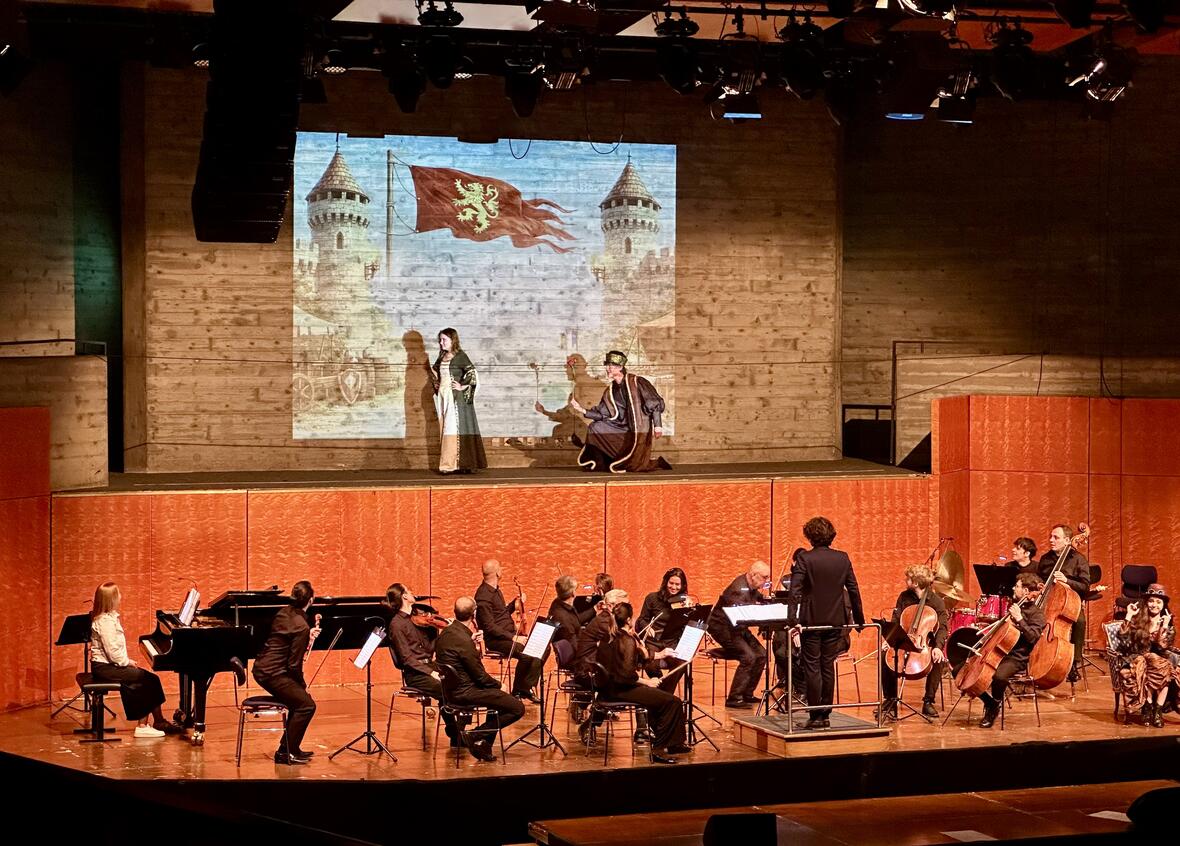 Szenenprobe mit zwei Schauspielern auf der Bühne und einem Orchester im Hintergrund.