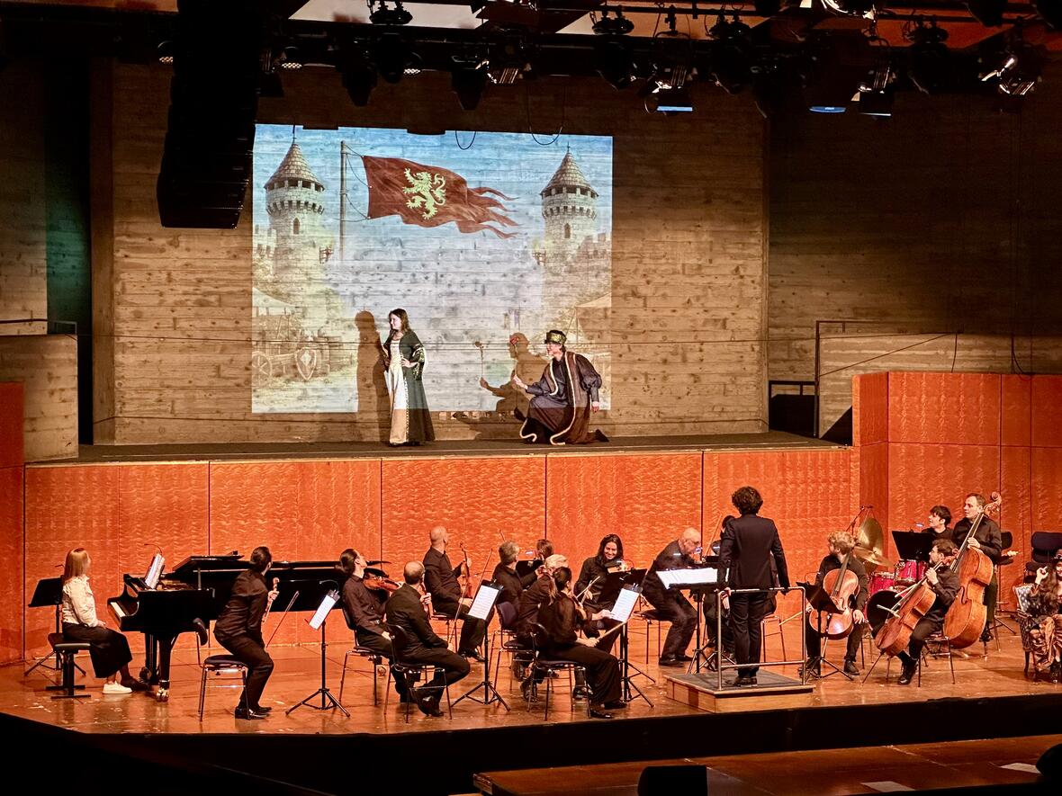 Szenenprobe mit zwei Schauspielern auf der Bühne und einem Orchester im Hintergrund.