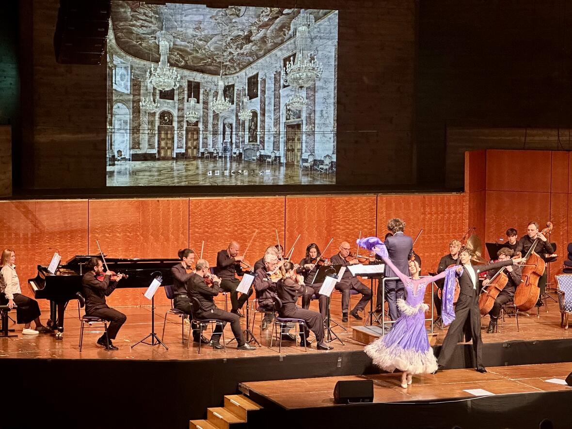 Tanzaufführung auf der Bühne mit Orchester, im Hintergrund historische Innenansicht.