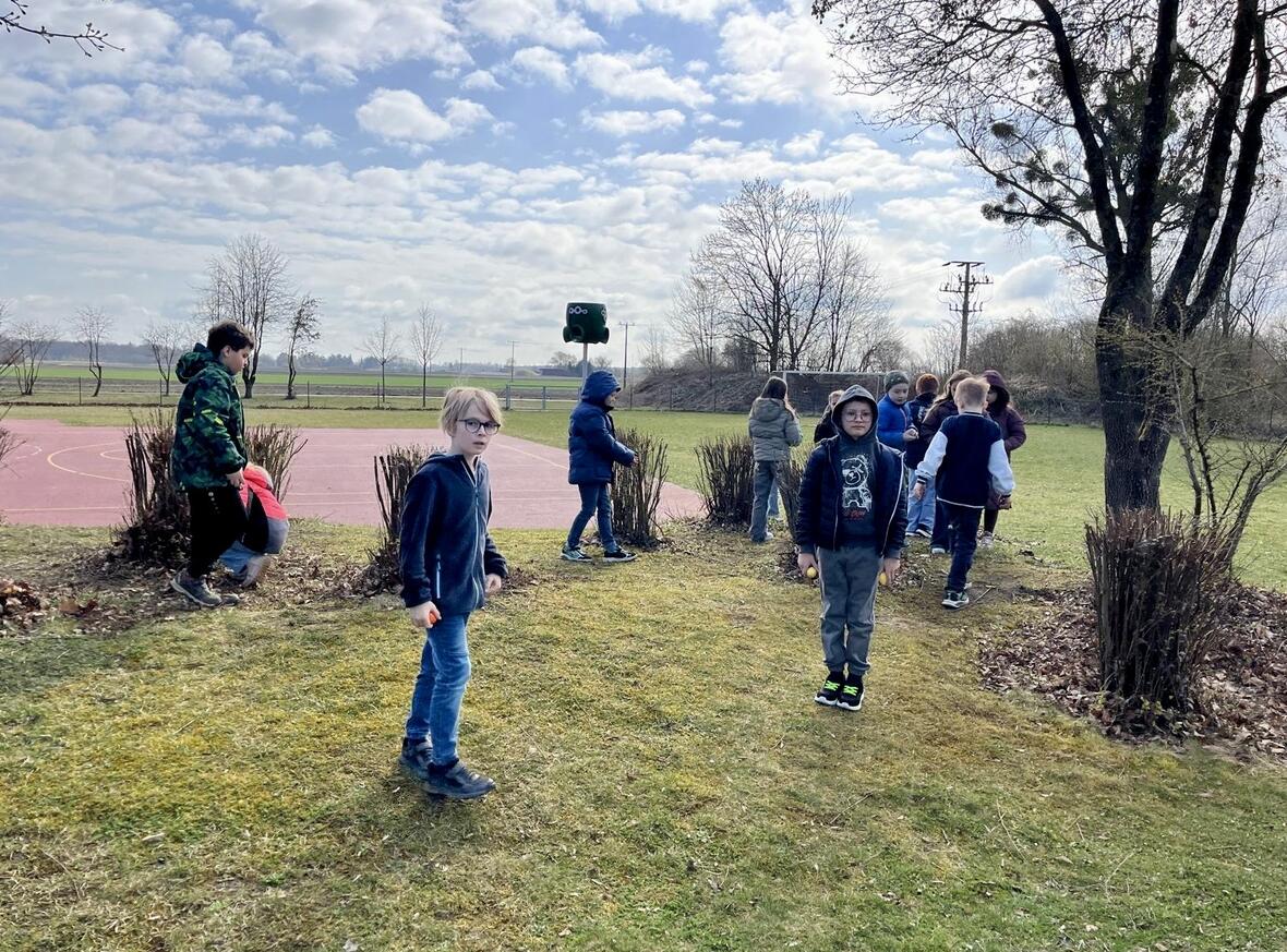 Kinder spielen auf einem Schulhof mit Basketballplatz, umgeben von Büschen und frischem Gras.