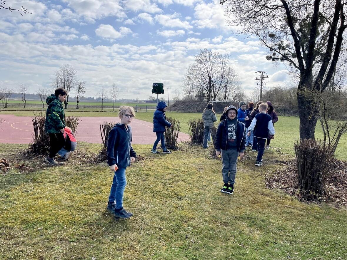 Kinder spielen auf einem Schulhof mit Basketballplatz, umgeben von Büschen und frischem Gras.