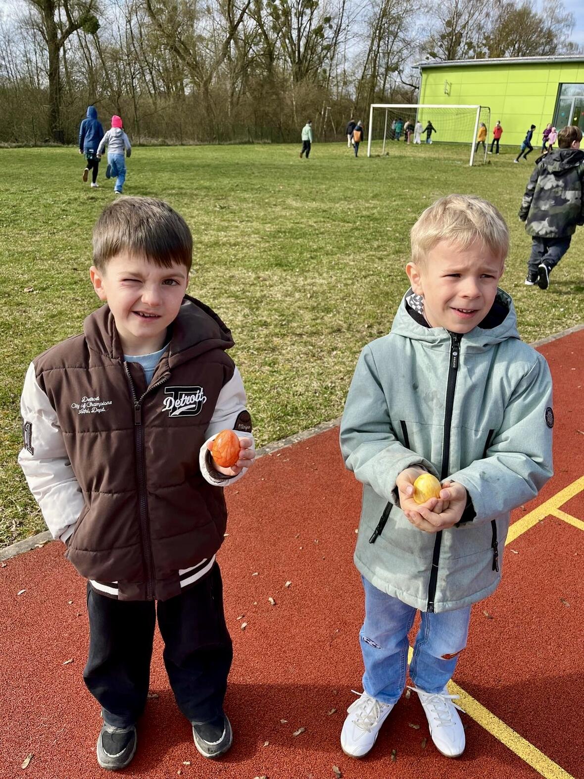 Zwei Kinder auf einem Sportplatz halten Obst in den Händen, während im Hintergrund andere Kinder spielen.