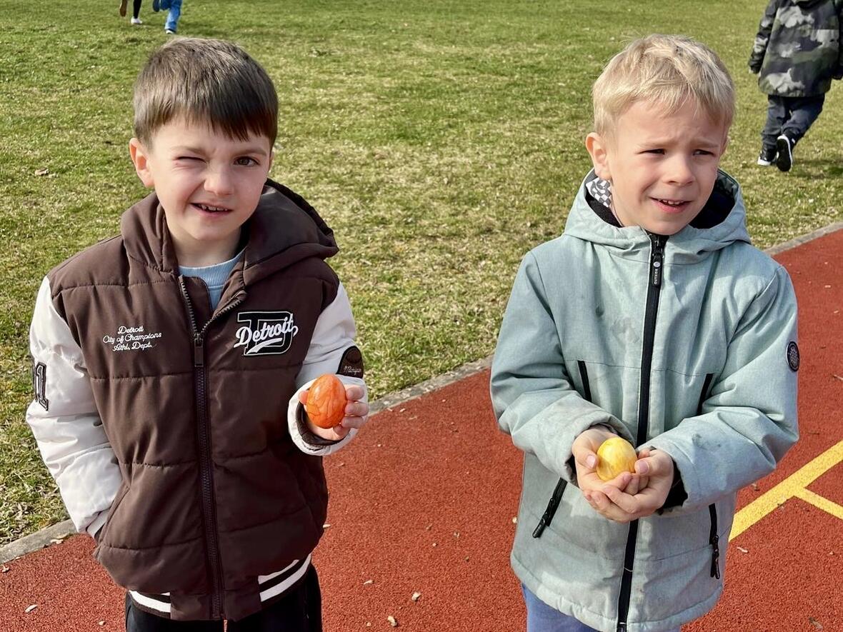 Zwei Kinder auf einem Sportplatz halten Obst in den Händen, während im Hintergrund andere Kinder spielen.