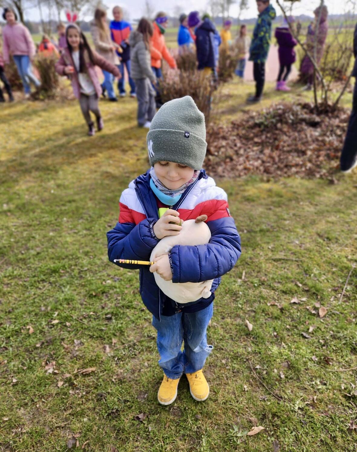 Junge mit Mütze und buntem Hoodie hält ein Stofftier und einen Bleistift, während im Hintergrund Kinder spielen.
