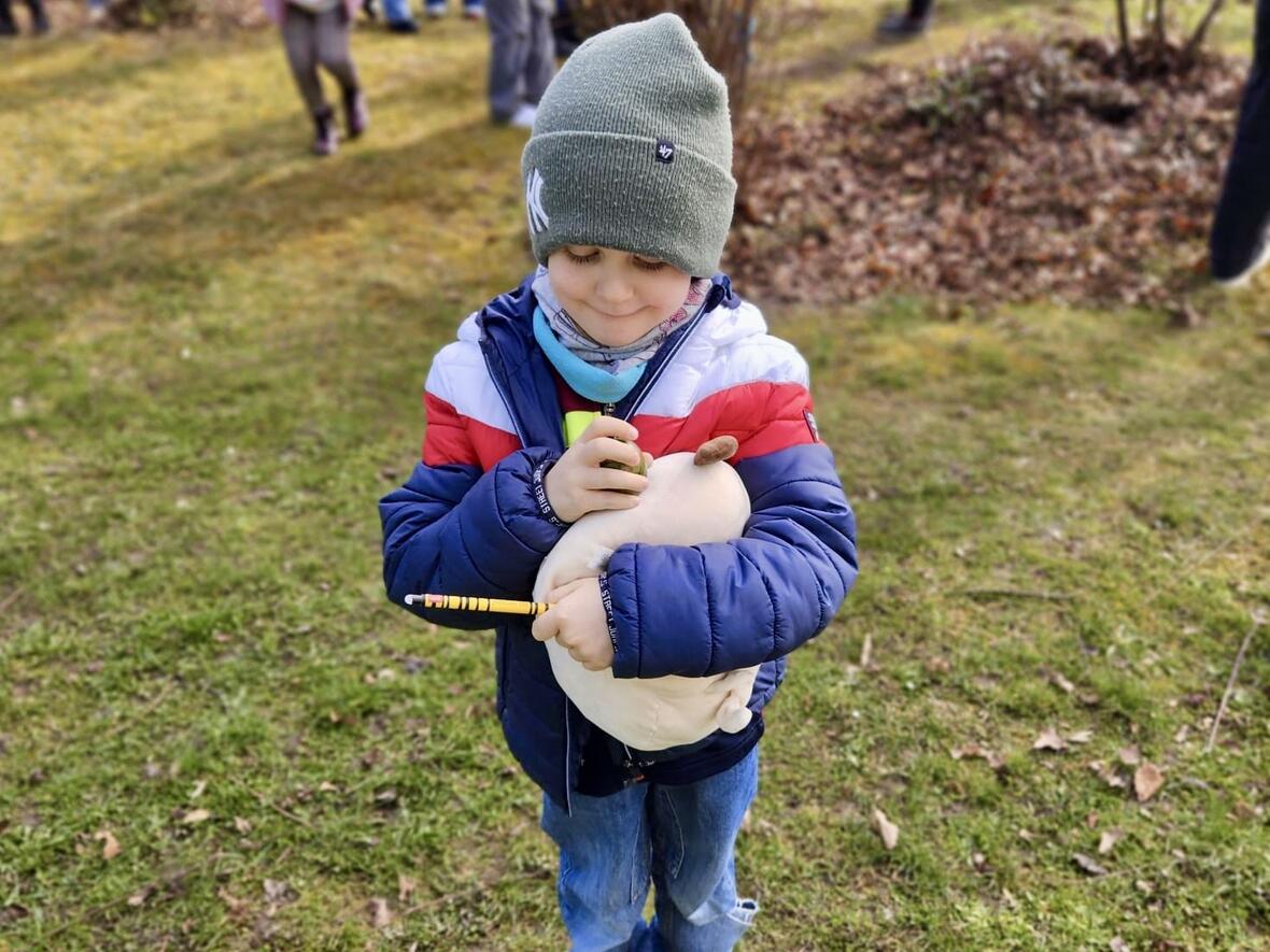 Junge mit Mütze und buntem Hoodie hält ein Stofftier und einen Bleistift, während im Hintergrund Kinder spielen.
