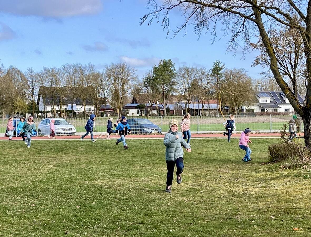 Kinder spielen und rennen fröhlich auf einer Wiese in der Nähe eines Sportplatzes.