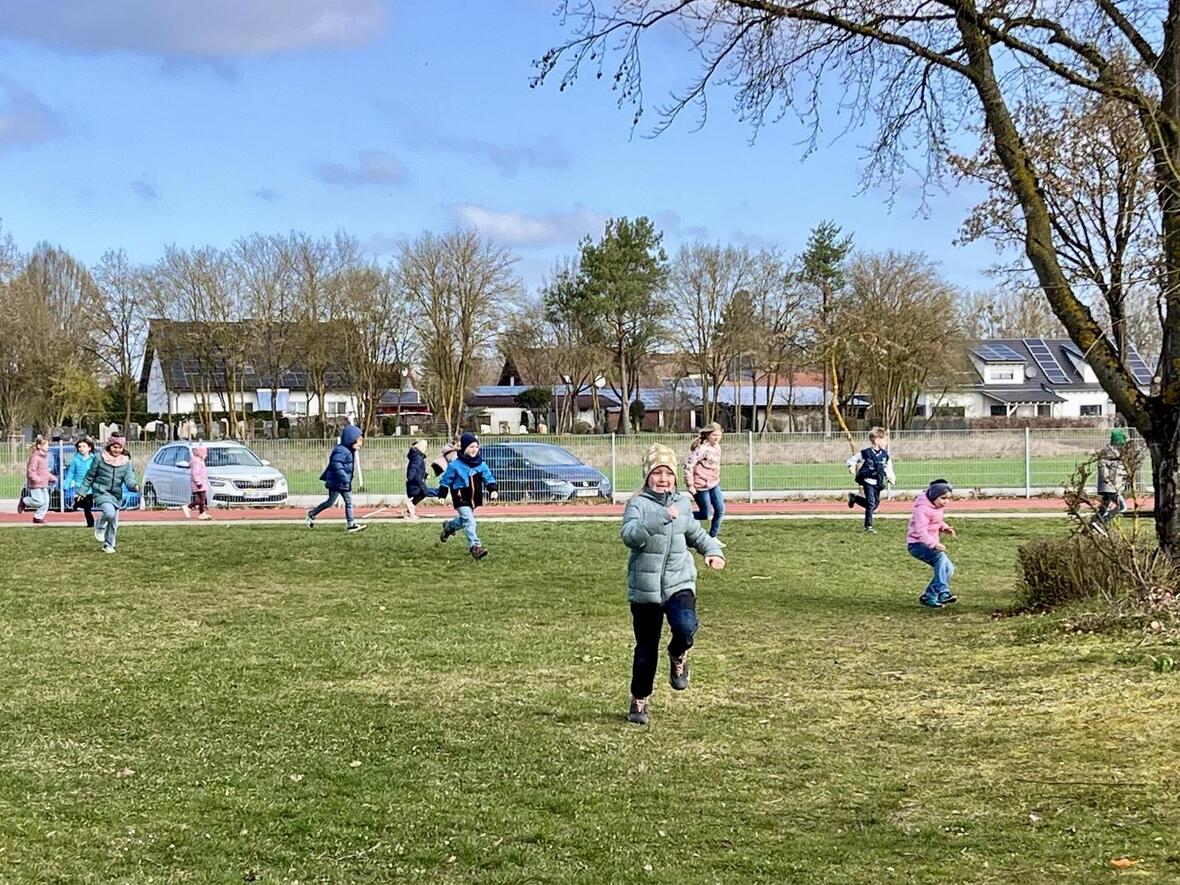Kinder spielen und rennen fröhlich auf einer Wiese in der Nähe eines Sportplatzes.